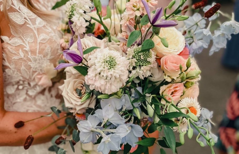 Braut hält einen bunten Brautstrauß mit Rosen, Astern und lila Blüten.