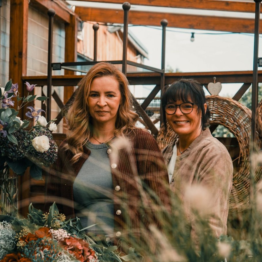 Zwei Frauen stehen entspannt inmitten von Blumen und Pflanzen auf einem romantischen Hintergrund.