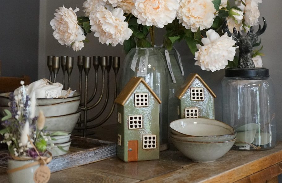 Zwei kleine Keramikhäuser, Blumen in Vasen und Schalen auf einem Holztisch.