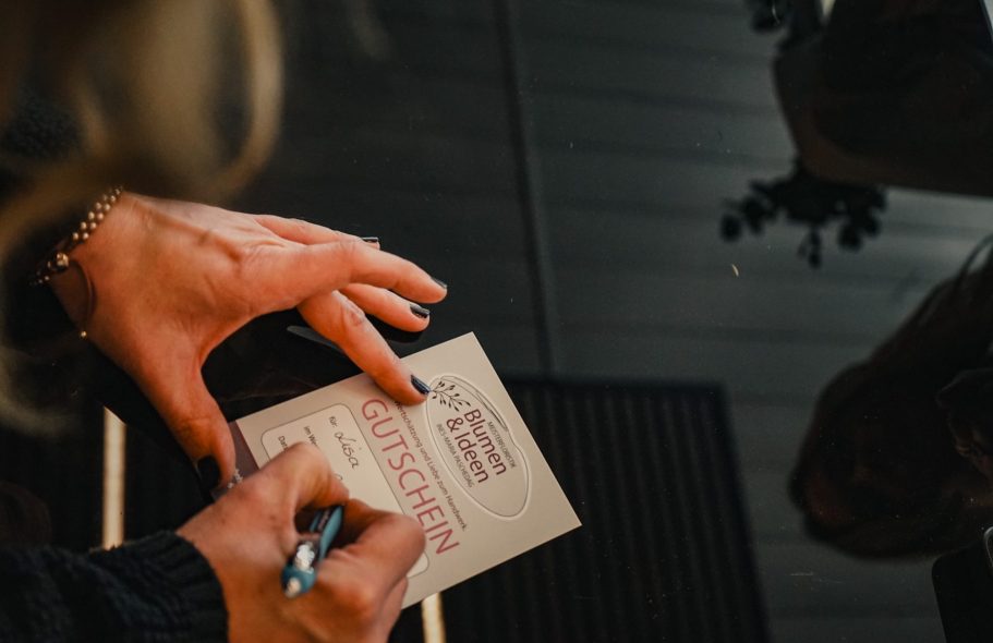 Eine Hand schreibt auf eine Gutscheinkarte, umgeben von einem Holzboden und Pflanzen.